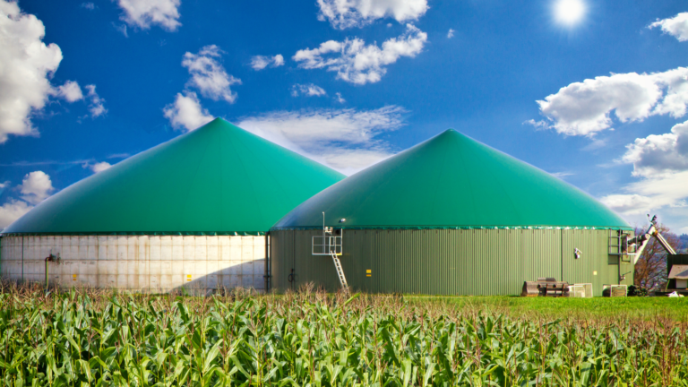 Instalações de biogás em área rural com tanques verdes e lavoura em primeiro plano, simbolizando produção sustentável de energia.