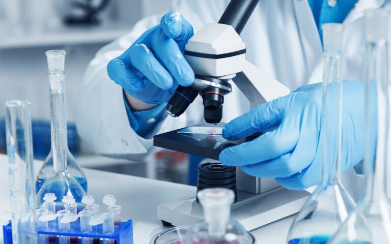 Pesquisador manuseando lâmina em microscópio com vidrarias de laboratório ao redor, representando investimento governamental em instituições científicas.