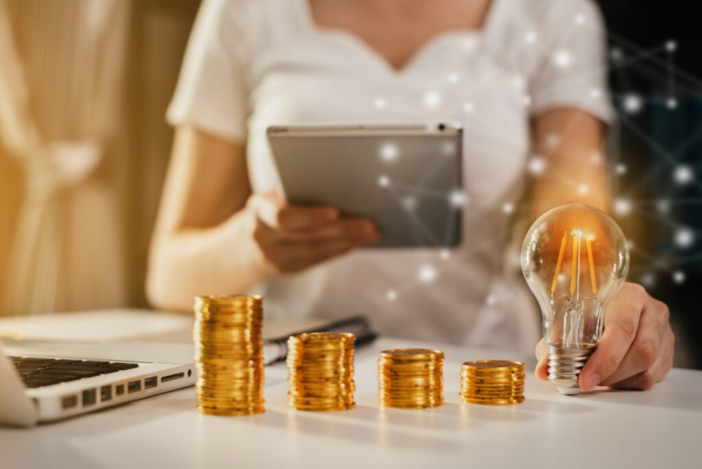 Pilhas de moedas e lâmpada simbolizando investimento, mulher usando tablet ao fundo — crédito recorde do BNDES de R$ 9 bilhões para inovação.