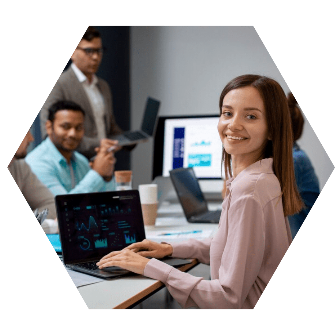Foto em formato hexagonal de um escritório: mulher sorrindo em primeiro plano olha para a câmera; ao fundo, colegas trabalham com notebooks e um monitor exibindo gráficos