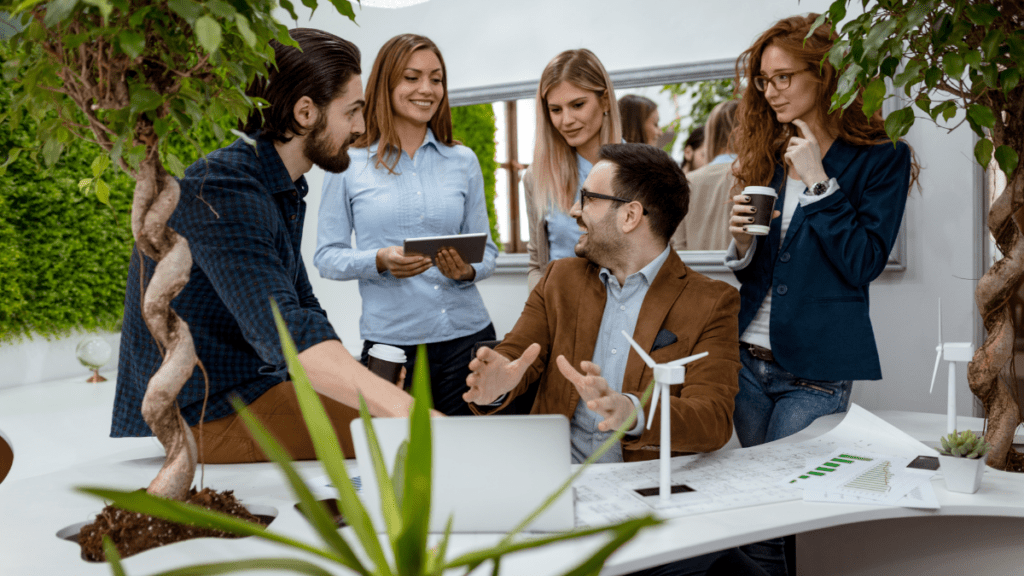 Equipe discutindo sustentabilidade em escritório verde, com mini turbina eólica na mesa e uma integrante usando tablet.