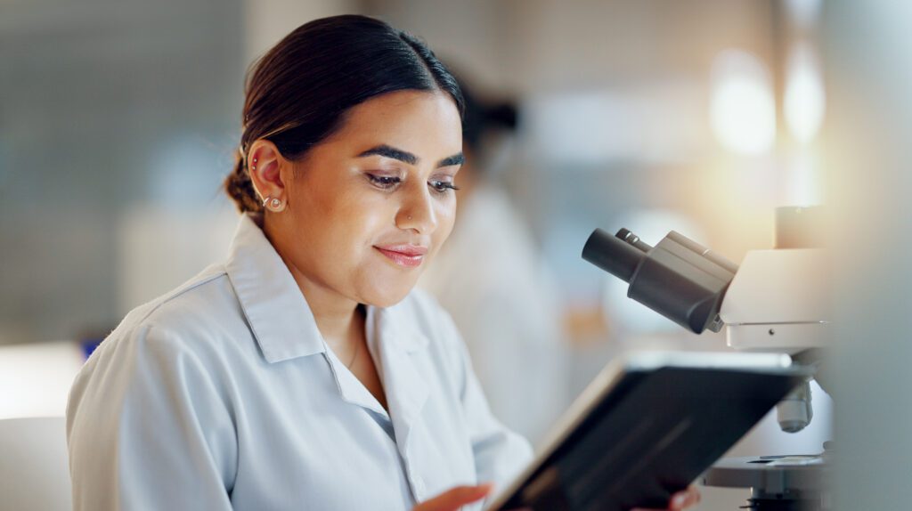 Pesquisadora de jaleco usa um tablet ao lado de um microscópio em laboratório, com fundo desfocado